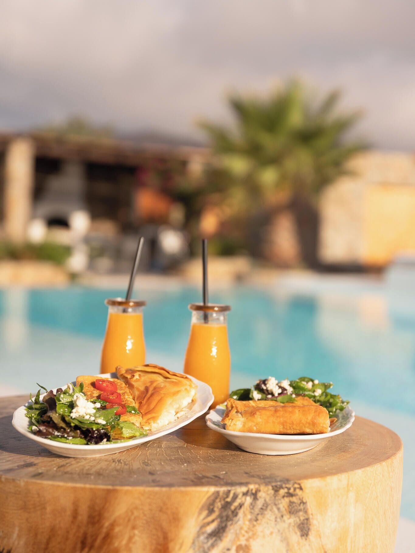 Mesa de madeira com pratos de comida e bebidas à beira da piscina.