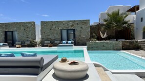 A pool area with lounge chairs, a stone wall, and a modern building with large windows. - Myconian Theros (Mykonos)