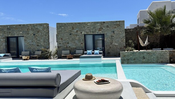 Un área de piscina con sillones de descanso, un muro de piedra y un edificio moderno con grandes ventanales.