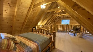 Une chambre confortable située dans les combles, avec un lit en bois, une vue sur l’extérieur par une fenêtre et une balustrade en bois entourant le balcon.