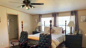 Ein Schlafzimmer mit einem Bett, zwei Stühlen, einem Nachttisch, einem Deckenventilator und einem Fenster mit Vorhängen.