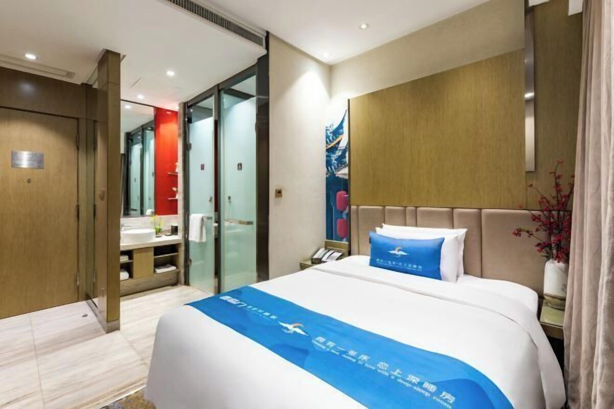 A hotel room with a bed, a nightstand, a mirror, a bathroom with a sink and toilet, and a door leading to another room.