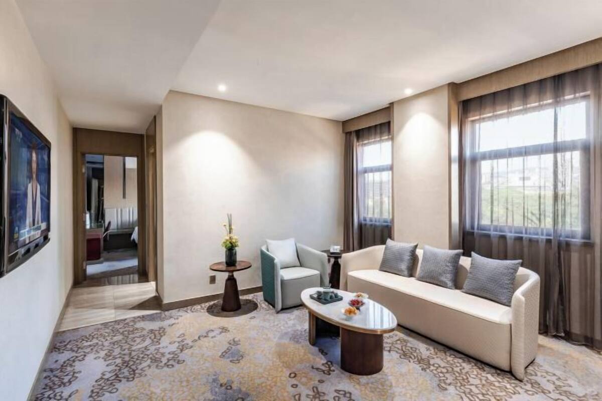 A modern hotel room with a sofa, a coffee table, and a small side table. There is a large window with curtains, a door leading to another room, and a patterned carpet.