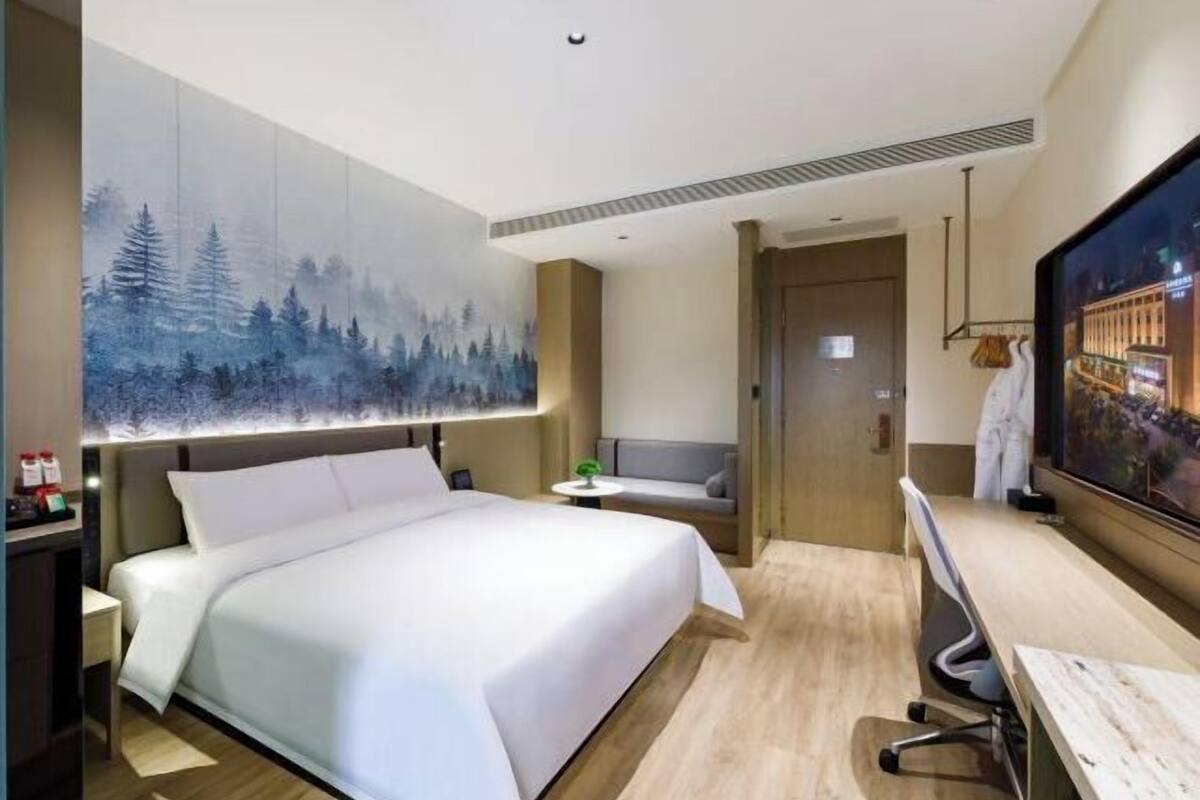 A modern hotel room with a large bed, a desk, a chair, a TV, and a painting of a forest on the wall.
