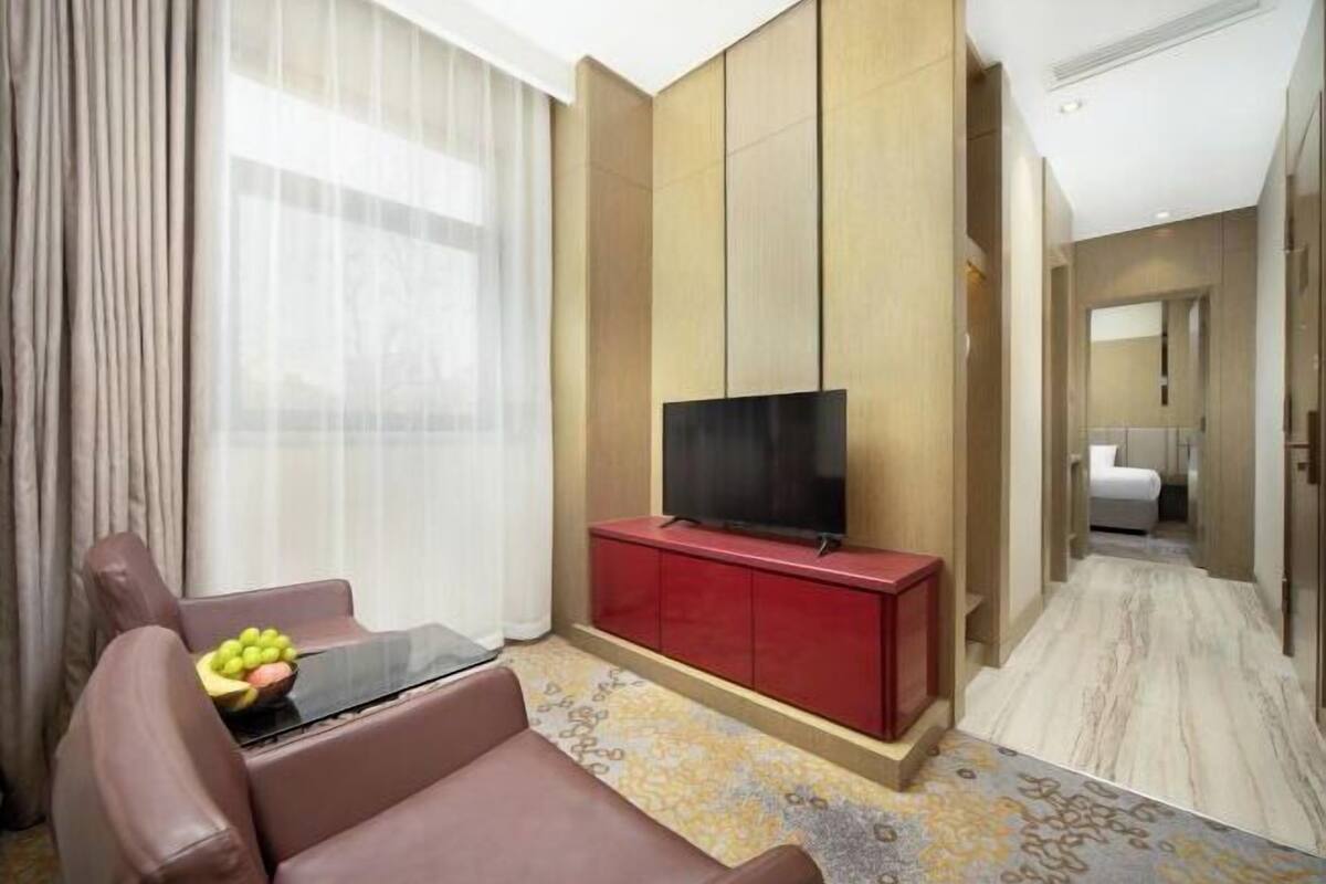 A modern hotel room with a leather sofa, a flat-screen TV on a red cabinet, a large window with sheer curtains, and a patterned carpet.