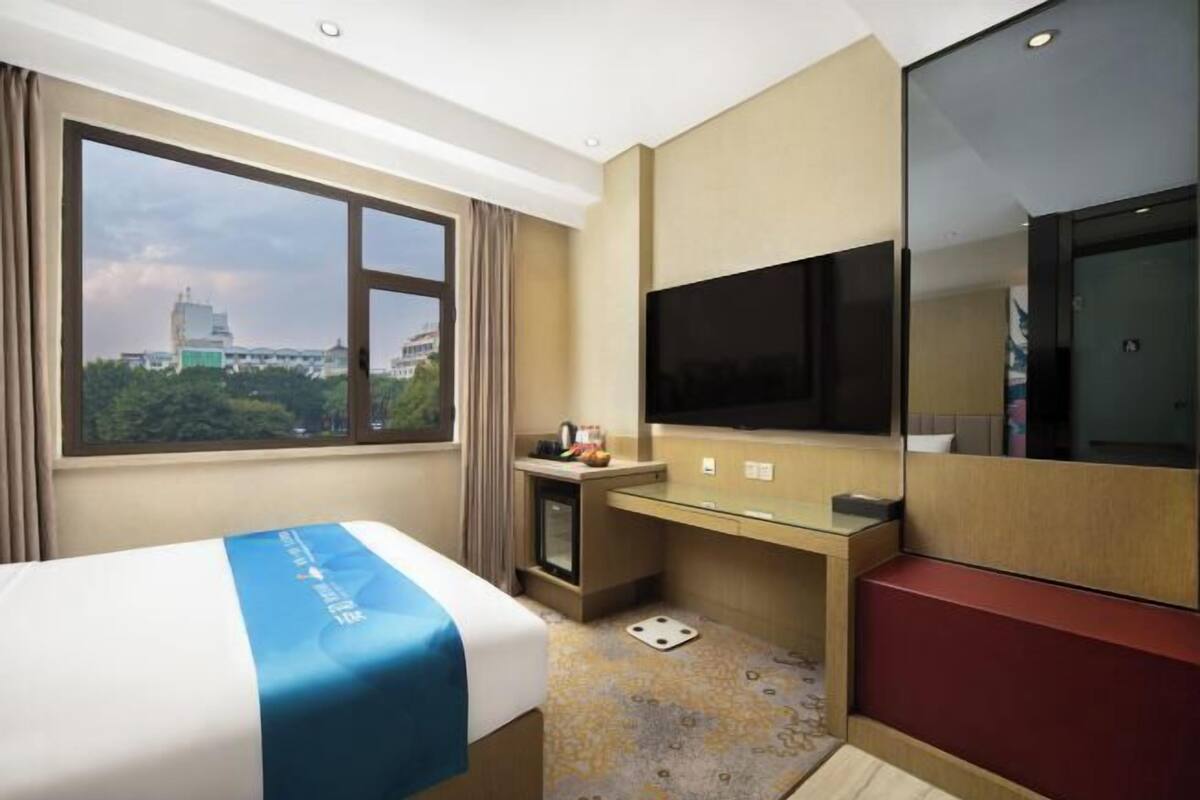 A hotel room with a bed, a desk, a television, and a large window offering a city view.