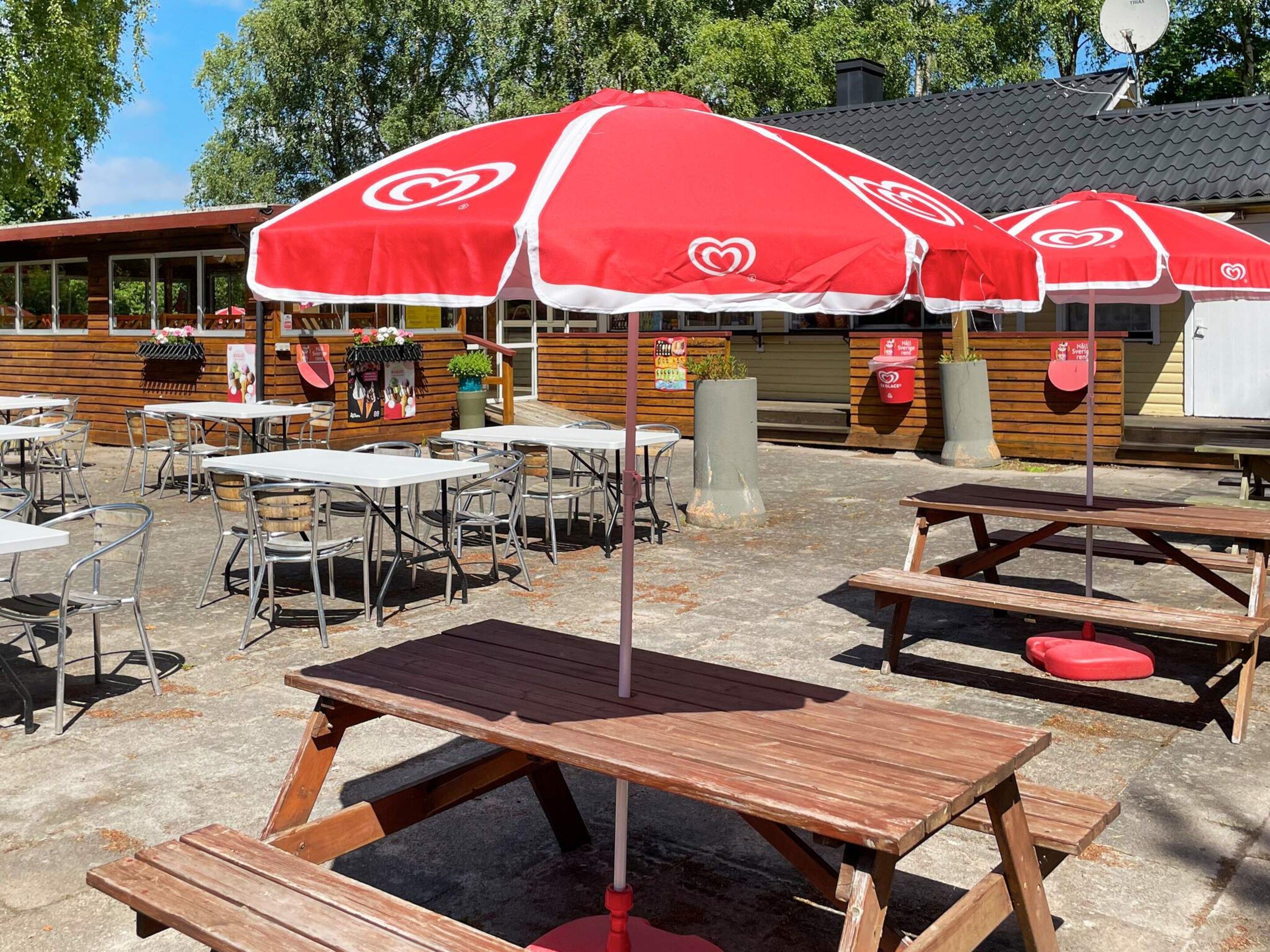 Outdoor tables and benches with red umbrellas, wooden fence, and a building with windows in the background.