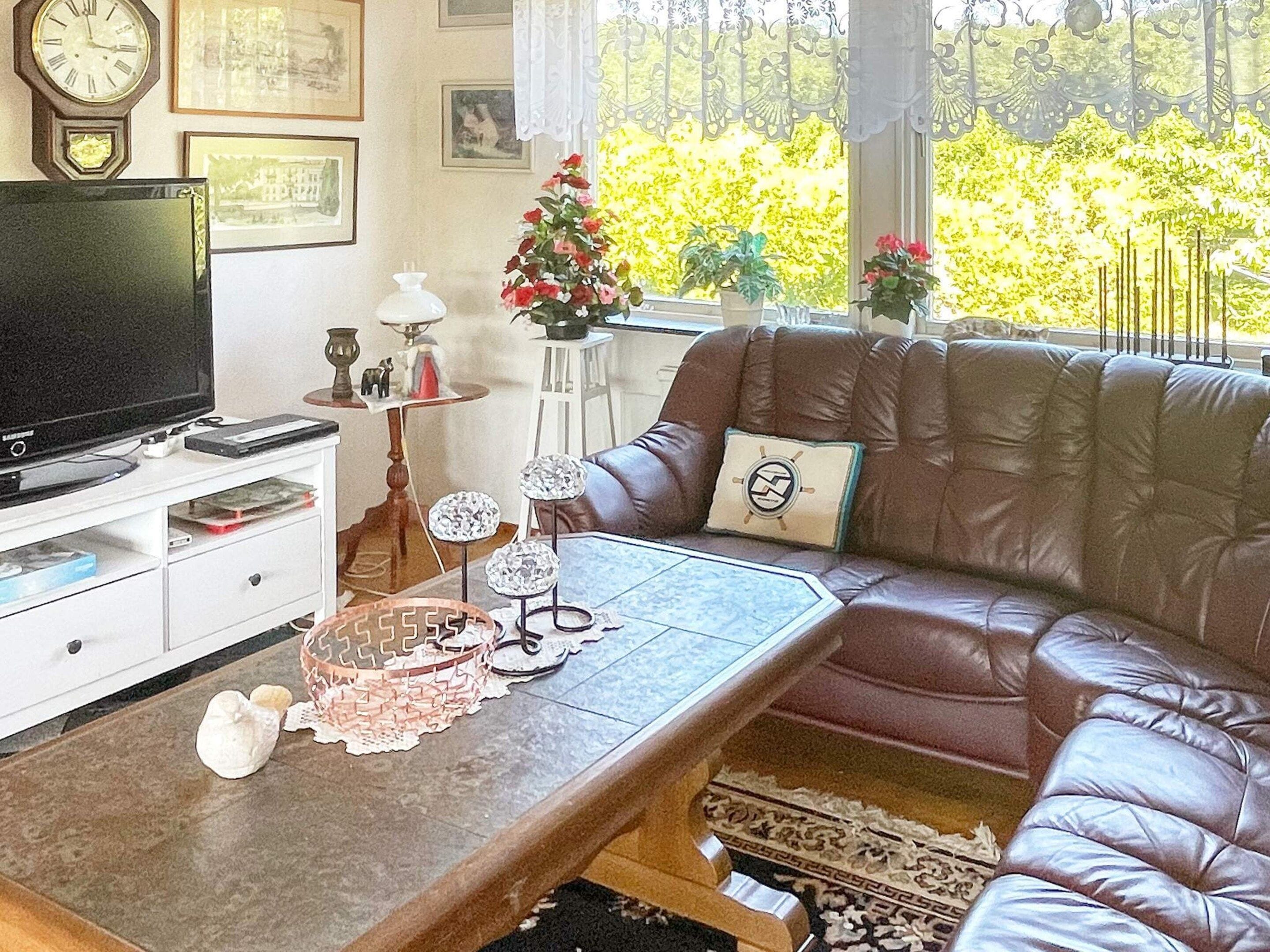 A living room with a leather sofa, a television on a stand, a coffee table with decorative items, and a view of greenery outside.