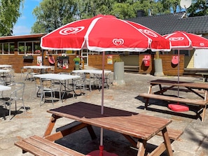 Outdoor tables and benches with red umbrellas, wooden fences, and a building with a sloped roof. - 4 Person Holiday Home in Solvesborg (Sölvesborg)