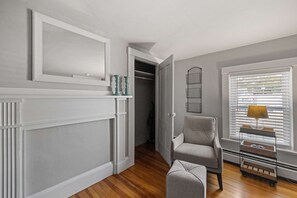 A hallway with a wooden floor, a grey armchair, a lamp on a cart, and a closet with a white door.
