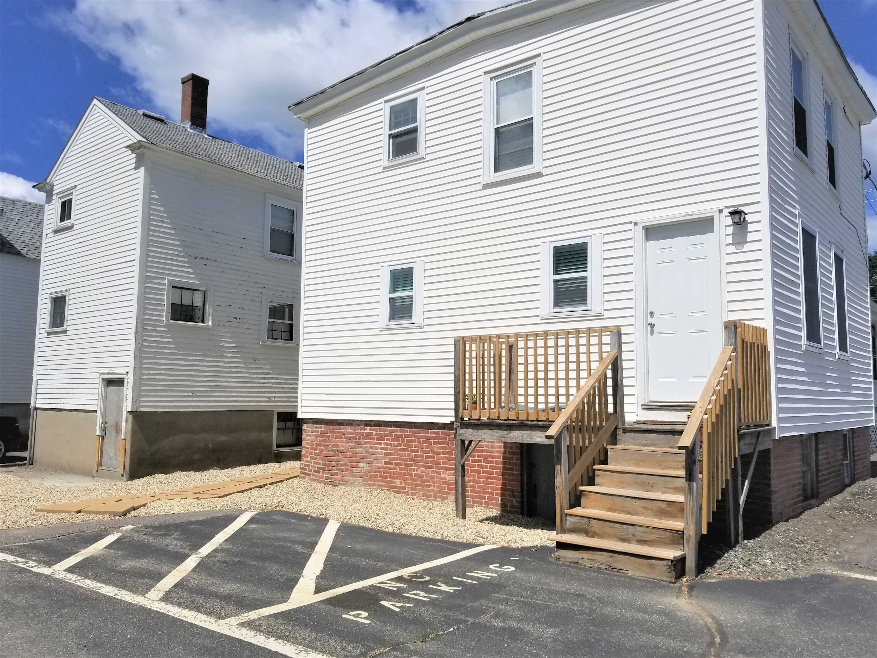 A two-story white building with a brick foundation, a wooden deck, and a parking area marked with "PARKING" on the pavement.
