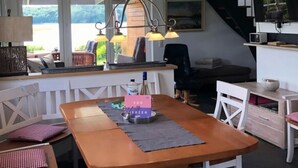 Una sala de estar acogedora con una mesa de comedor de madera, sillas blancas, una escalera y un amplio ventanal con vistas al verdor.