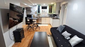 Ein kompakter Wohnbereich mit Sofa, Couchtisch, Wand-Fernseher, Küchenzeile mit Schränken und Mikrowelle sowie Esstisch mit Stühlen.
