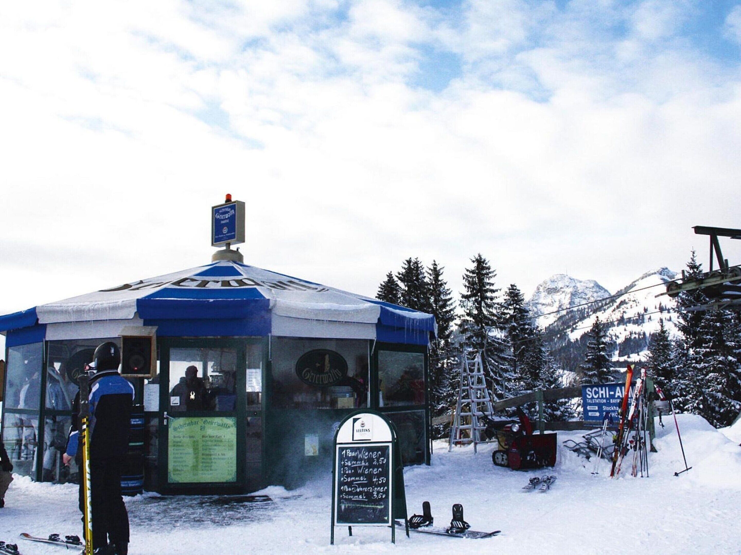 A snow-covered mountain area with a small building, likely a ski lodge, featuring a glass front and a signboard with prices.