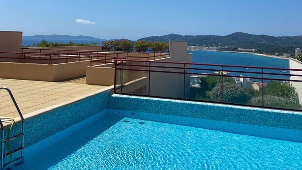 A rooftop swimming pool with clear blue water, surrounded by a red railing, and offering a view of the sea and distant hills.
