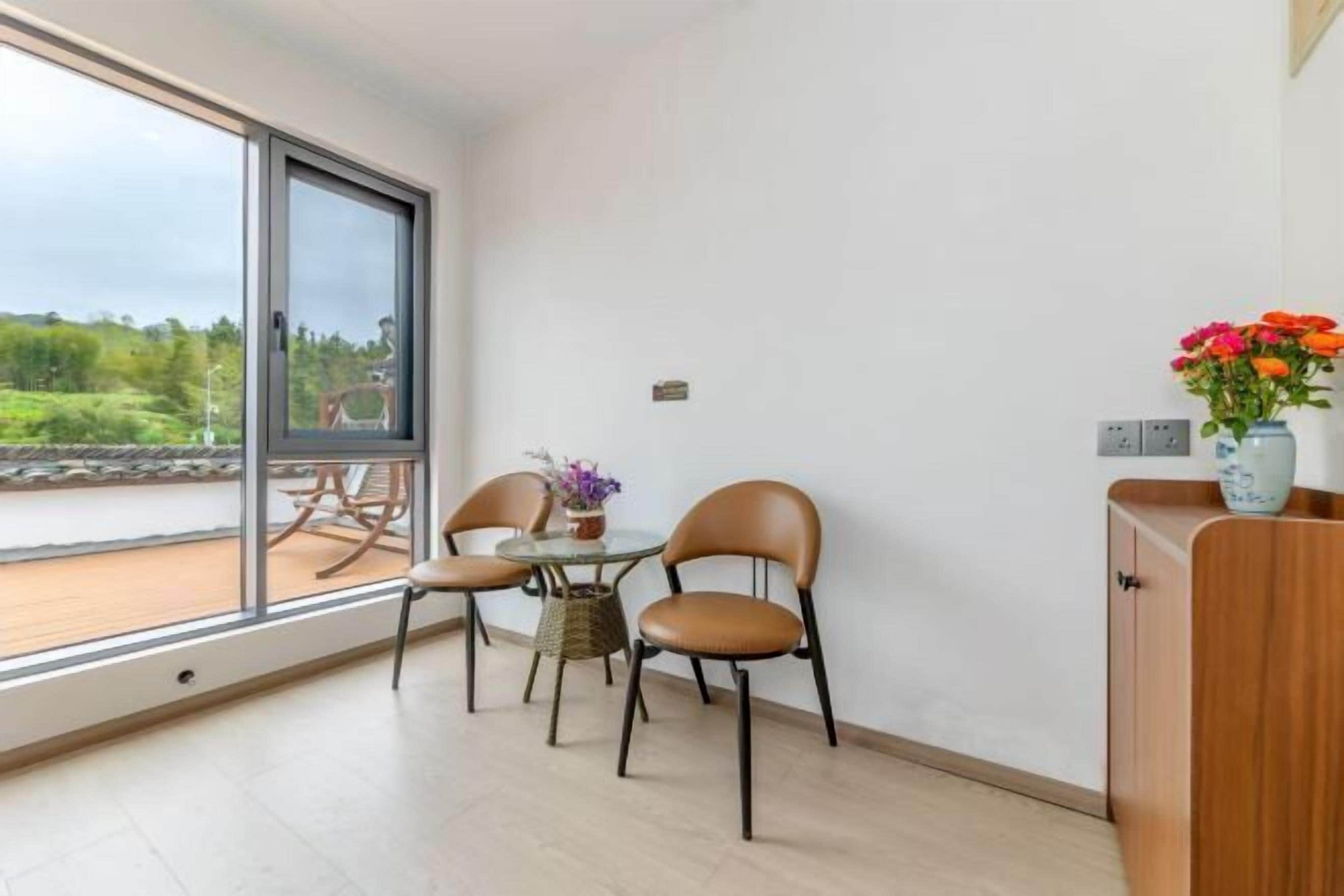 Una habitación con un ventanal grande, una mesa con sillas, un jarrón pequeño con flores y un armario de madera.