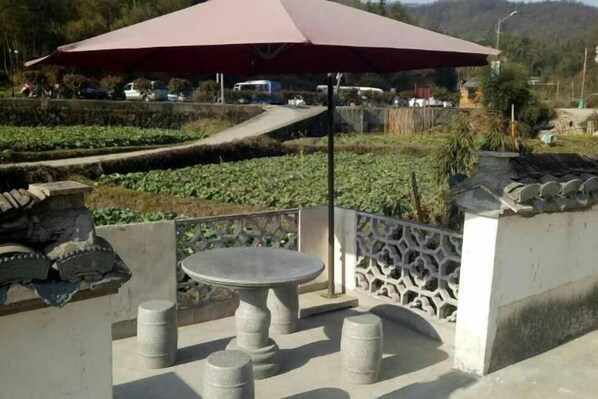 Una terraza con una mesa y taburetes, una cerca decorativa, y vistas a un área ajardinada con una carretera y vehículos al fondo.