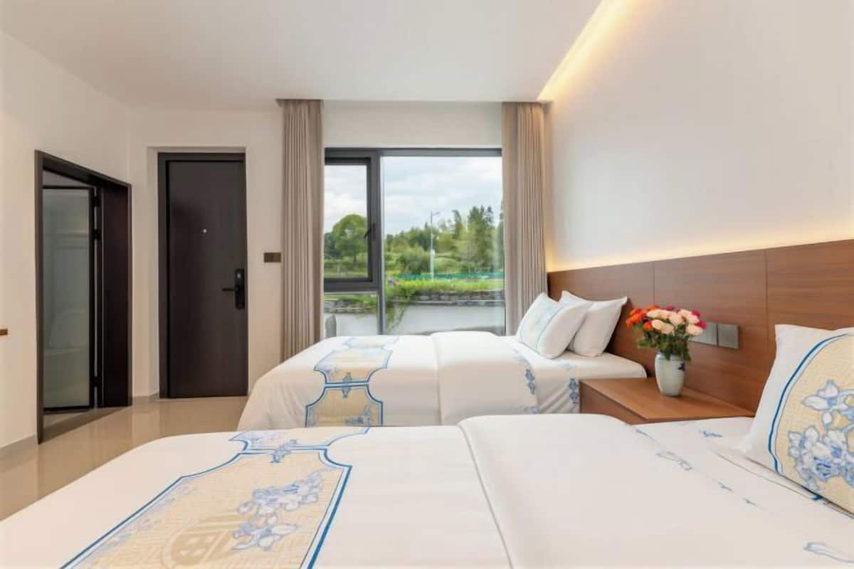 Une chambre d’hôtel avec deux lits, une grande fenêtre, une porte et une table de chevet ornée d’un vase de fleurs.