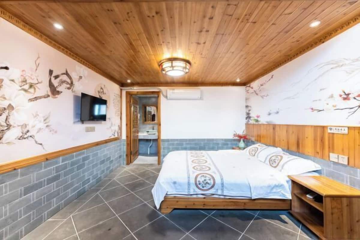 Une chambre avec un plafond en bois, un lit recouvert d’une couvre-lit à motifs et un téléviseur fixé au mur.