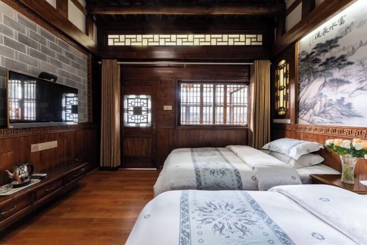 Una habitación tradicional con paredes de madera, una pared de ladrillo, un televisor, una cama con ropa de cama estampada, un escritorio con una tetera y un cuadro en la pared.