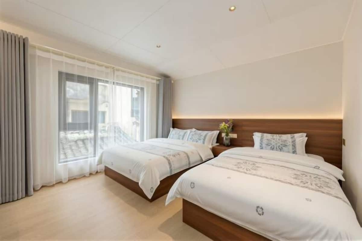 Une chambre d’hôtel avec deux lits, une grande fenêtre avec des rideaux, une tête de lit en bois et une petite table de chevet surmontée d’un vase.