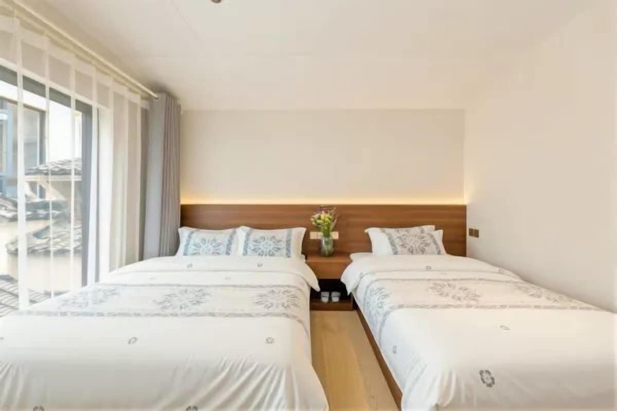 Habitación de hotel con dos camas, una mesita de noche con un florero y vistas al exterior.