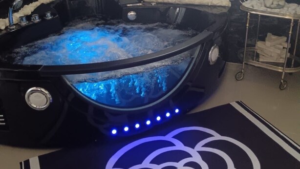 Ein Wellnessbereich mit Whirlpool, umgeben von weißen Badteppichen, einer Teppichauflage mit Rosenmuster und einem kleinen Tisch mit Badeutensilien.