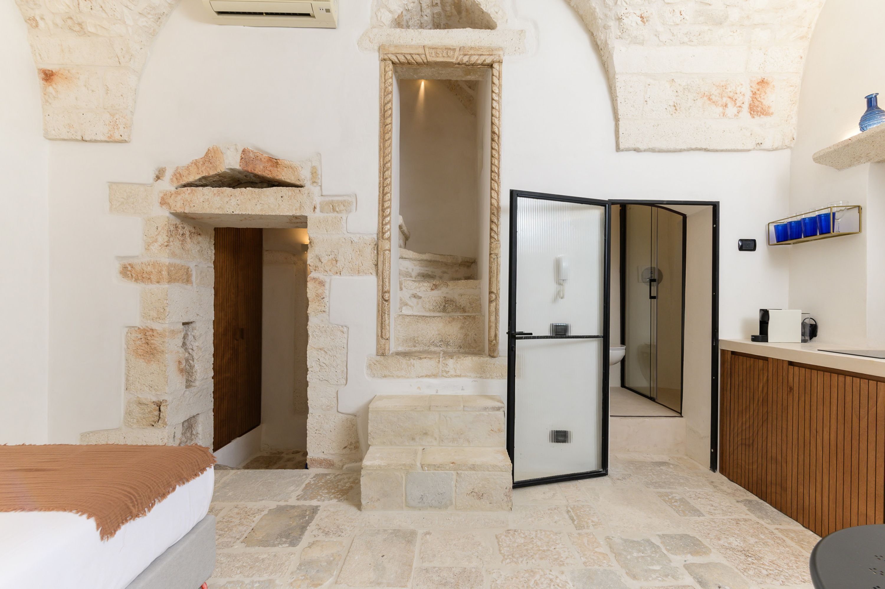 A compact room with a stone wall, a bed, a built-in bench, a small kitchen area, and a door leading to a staircase.