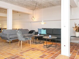 Una sala de estar moderna con un sofá gris, sillones y una mesita de centro. Hay lámparas colgantes y un jarrón con flores sobre una mesita auxiliar.