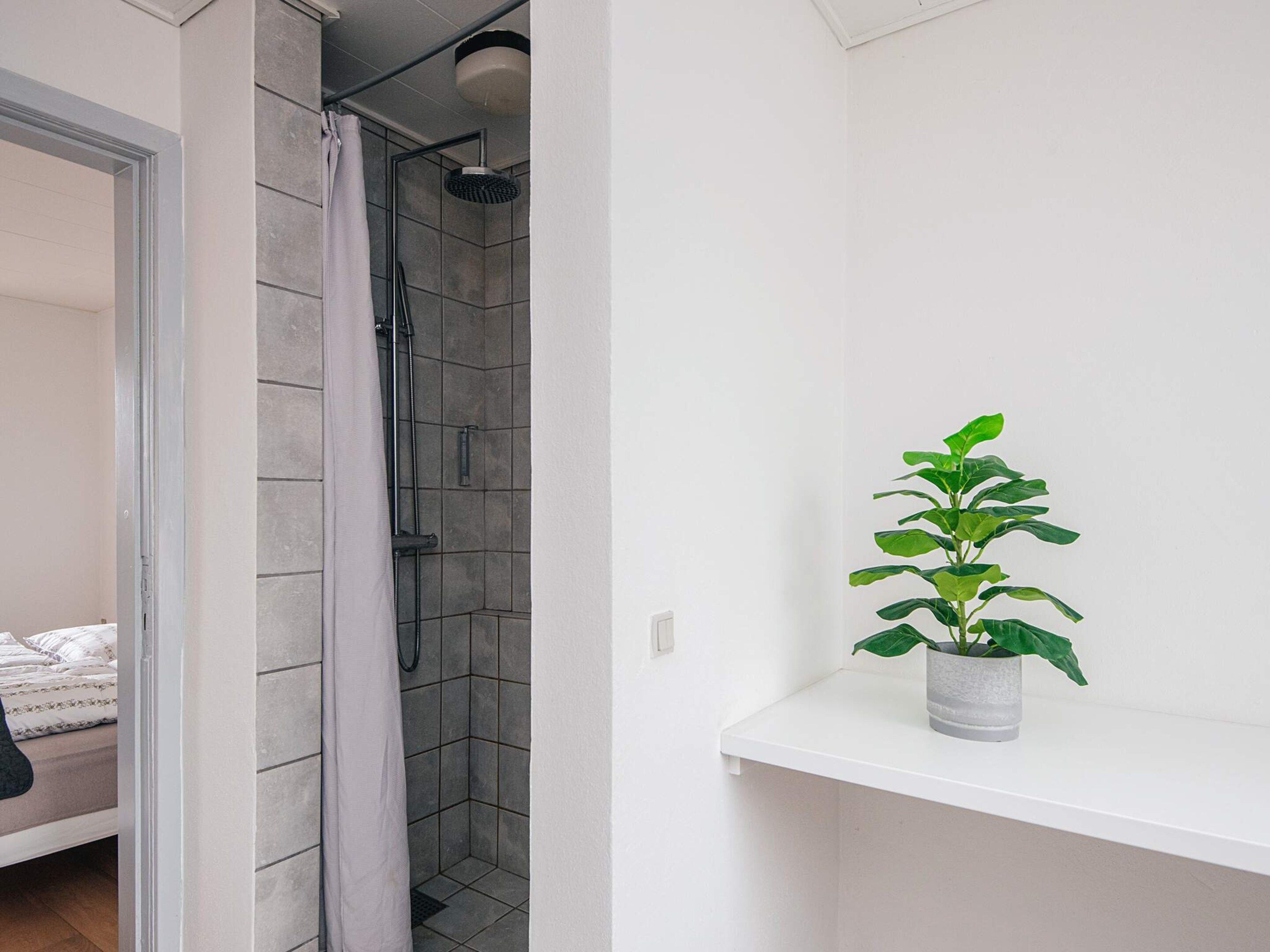 Un baño con ducha, una planta en maceta sobre una estantería y una cama en la habitación contigua.