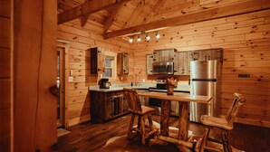 A rustic cabin interior with wooden walls, a wooden dining table, chairs, a refrigerator, a microwave, and a sink.