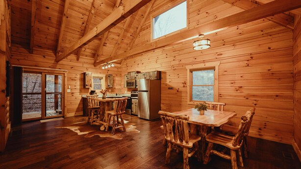 A wooden cabin interior with a dining area, kitchen, and large windows.