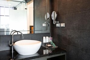 Une salle de bain moderne avec un grand lavabo sur pied blanc, des murs recouverts de carreaux foncés, un miroir avec deux appliques murales et une petite étagère contenant des articles de toilette.
