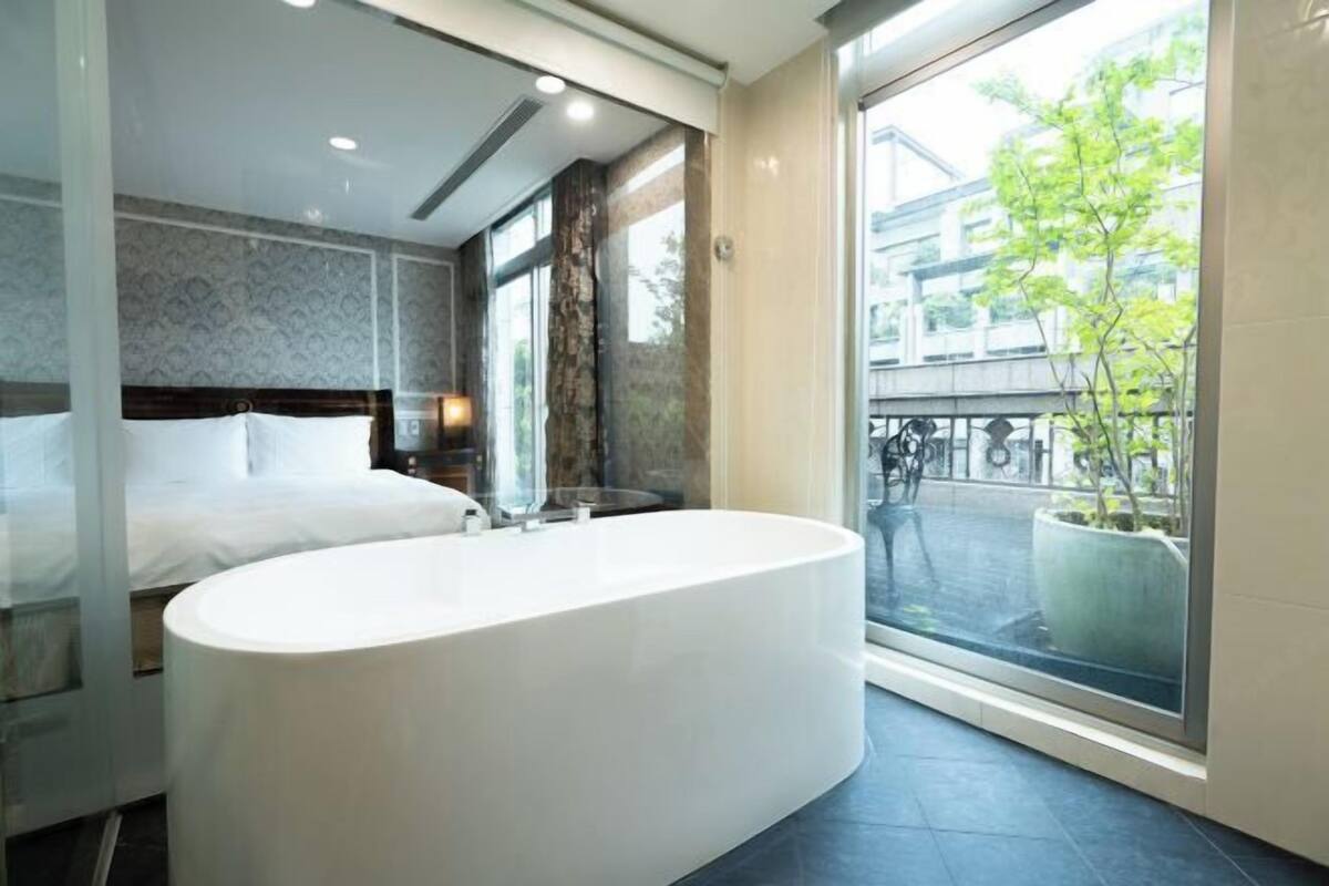 Un baño moderno con una bañera independiente, un espejo grande y vistas a un paisaje urbano a través de un ventanal.