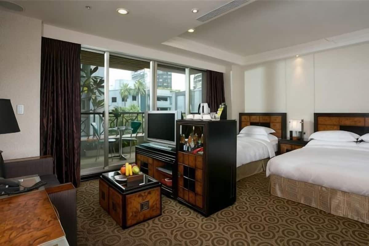 Habitación de hotel con cama, televisión, escritorio con computadora, mesa de centro con fruta y un amplio ventanal con cortinas.