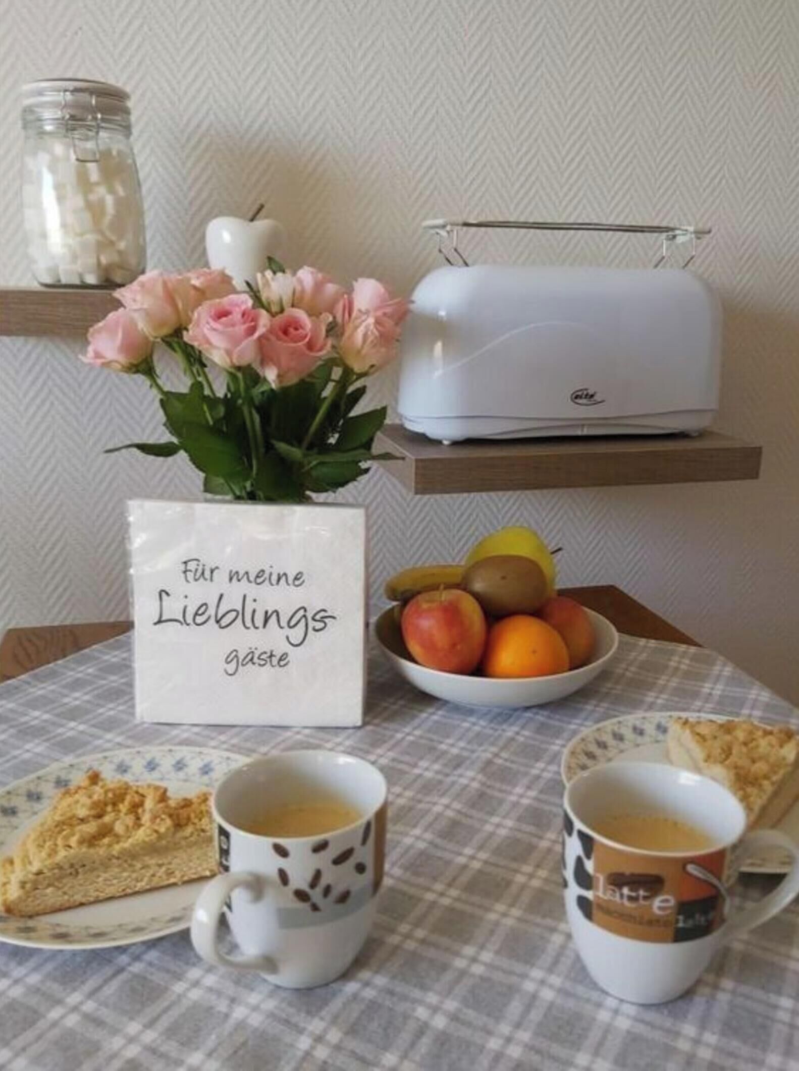 A breakfast setting with a cake, coffee cups, a fruit bowl, a toaster, and a vase with flowers.