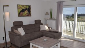 A modern living room with a sectional sofa, a coffee table, a floor lamp, a painting, a lamp on a side table, and a view of a deck through a glass door. - Holiday Apartment Falkenlay (Bad Bertrich)