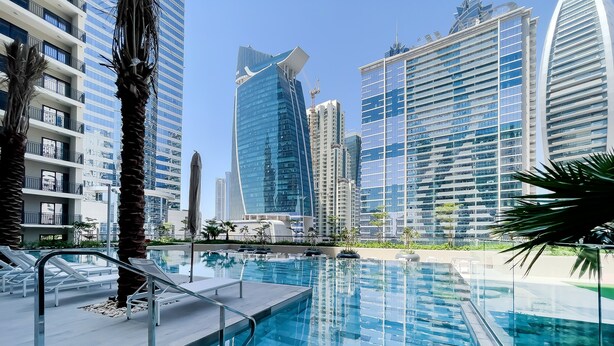 A modern hotel pool area with clear blue water, surrounded by high-rise buildings and palm trees. - Zada Tower 1BR (Dubai)