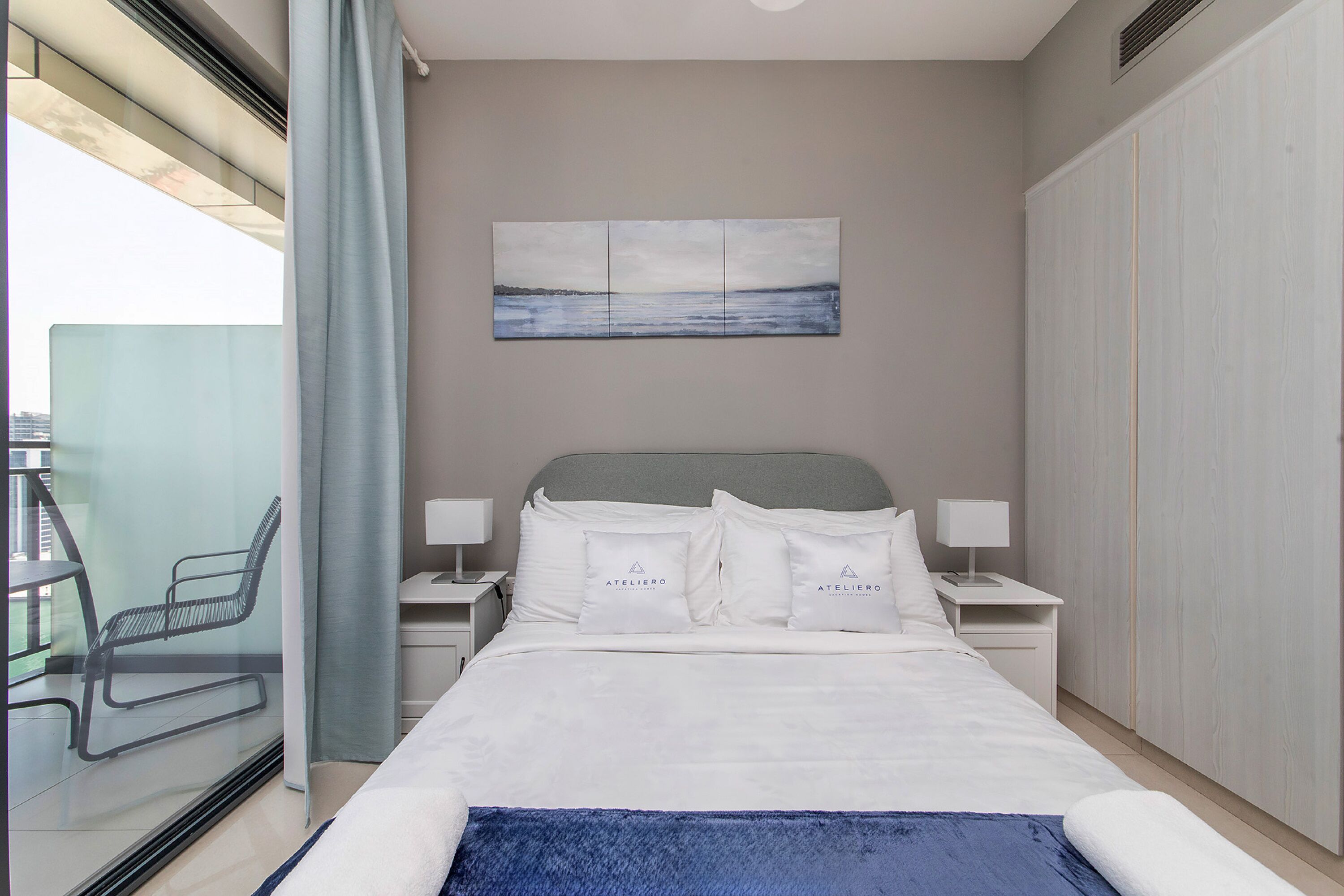 A hotel room with a bed, two bedside tables, a nightstand, a chair, a window with a view, and a painting on the wall.
