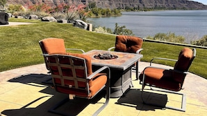 Un patio exterior con una mesa y sillas alrededor de un pozo de fuego, con vistas a un lago y montañas.