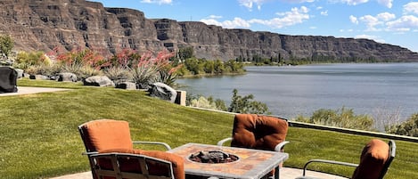 An outdoor patio with a fire pit table and chairs, overlooking a lake and mountains.