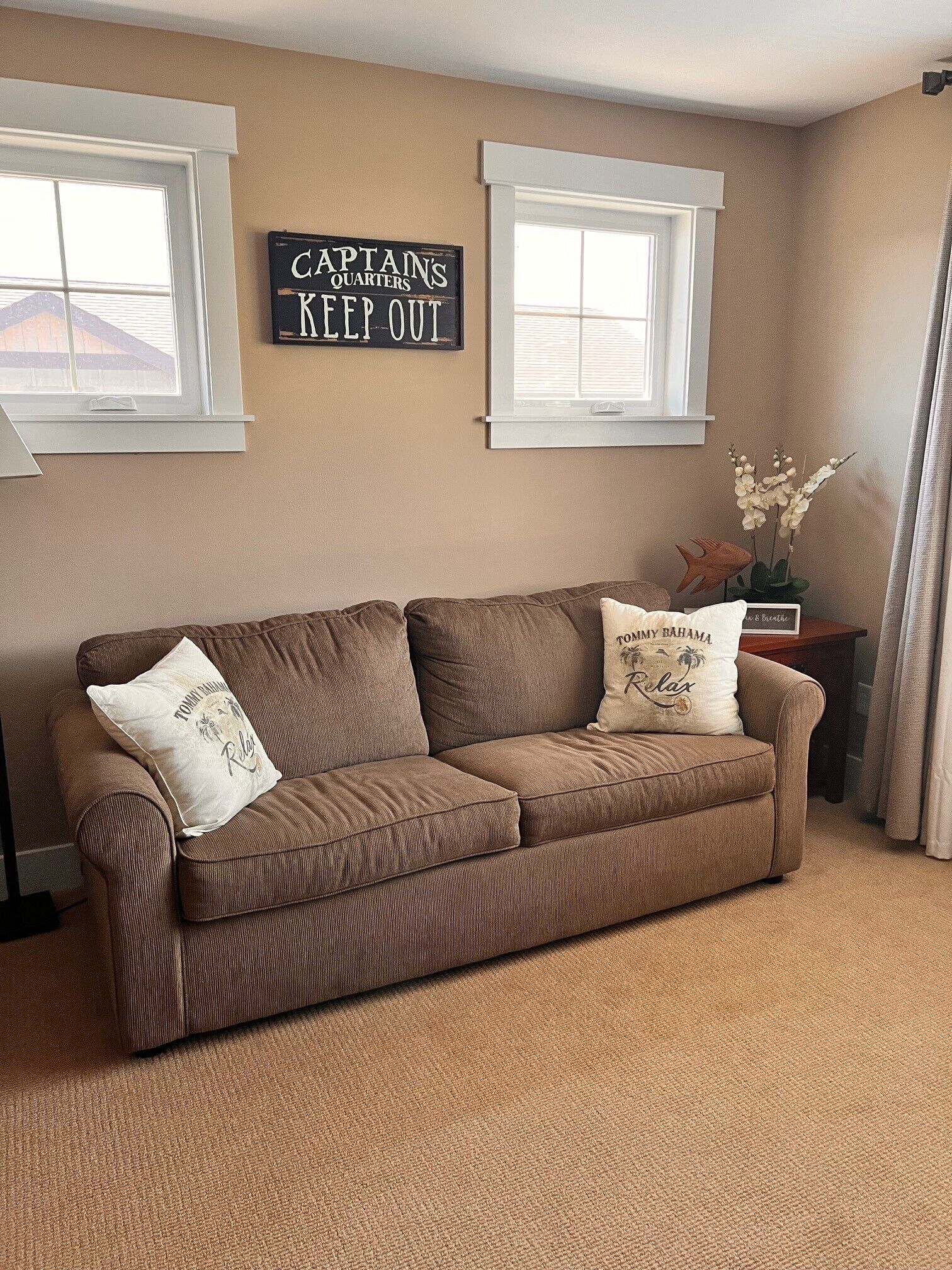 A brown sofa with two decorative pillows, a wall-mounted sign, and a small table with a vase of flowers.