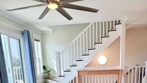 Una sala de estar con un sofá color beige, un ventilador de techo y una escalera que sube a un nivel superior.
