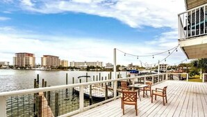 Una terraza de madera con mobiliario exterior, con vista a una masa de agua y edificios al fondo.