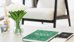 Ein sauberer, moderner Schreibtisch mit einem grünen Buch mit dem Titel "The Rolex Book", einem schwarzen Buch und einer klaren Vase mit grünen Blättern.