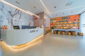 A reception area with a white counter, a computer, and a wall with a tree design. There are shelves with various items and a display case with products. - Dali Teng'ange Smart Hotel (Dali High-speed Railway Station) (Dali City)