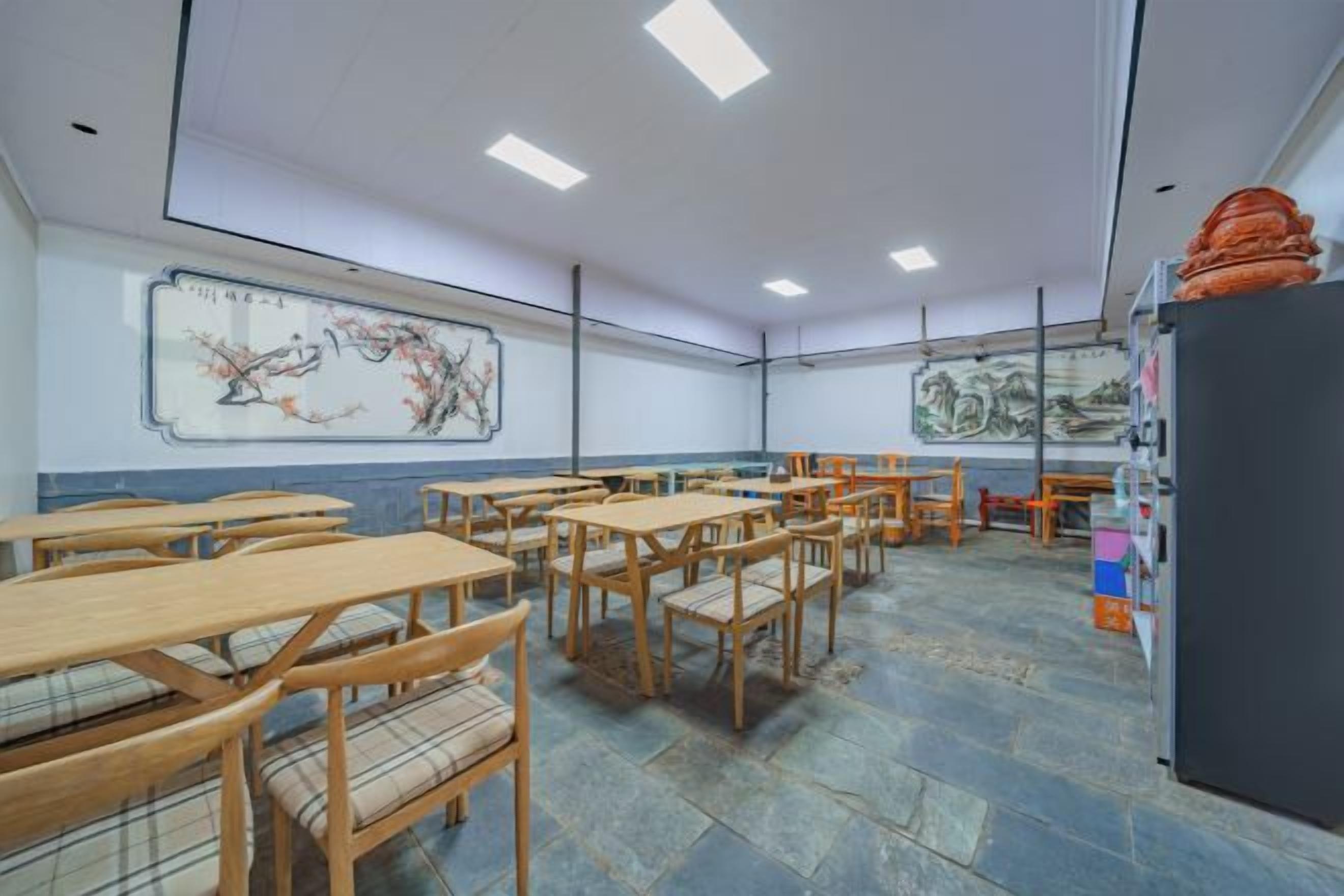 A classroom with wooden desks and chairs, a blackboard with a painting, and a refrigerator on the right.