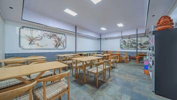 A classroom with wooden desks and chairs, a blackboard with a painting, and a refrigerator on the right.