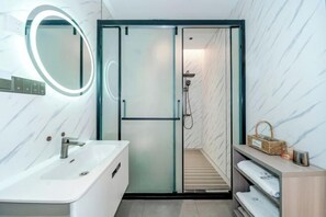 A modern bathroom with a vanity, a mirror with a light, a shower area, and a shelf with towels. - Dali Teng'ange Smart Hotel (Dali High-speed Railway Station) (Dali City)