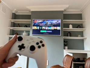 A living room with a TV displaying Xbox games, a white game controller, and shelves with decorative items.